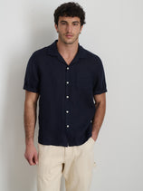 A man with short curly hair wears the Camp Shirt In Linen, a dark navy men's short-sleeve shirt, paired with light beige pants. He stands against a white background with one hand in his pocket.