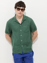 A person stands against a plain white background with their hands behind their back, wearing sunglasses, blue shorts, and the Camp Shirt In Linen in green with short sleeves.