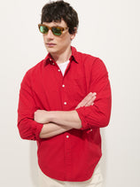 A person wearing a bright red Mill Shirt in Cotton Poplin over a white crewneck t-shirt and tortoise-shell sunglasses with green lenses, standing with their arms crossed.