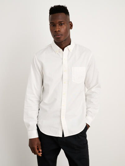 A man stands against a plain white background, wearing the Mill Shirt in Cotton Poplin with French seams and real shell buttons, paired with black pants and one hand in his pocket.