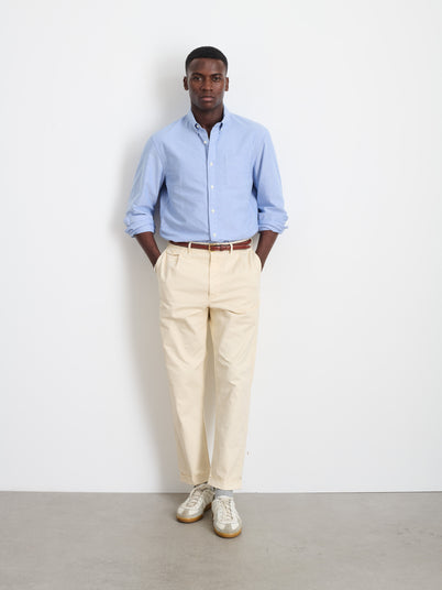 A man stands against a white wall wearing a light blue button-down shirt, Standard Pleated Pant in Chino (beige), and white sneakers, with his hands in his pockets.
