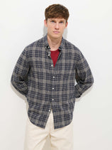 A young man with short brown hair wears the Mill Check Shirt In Linen Cotton over a red T-shirt and off-white pants, standing against a plain white background with his hands behind his back.