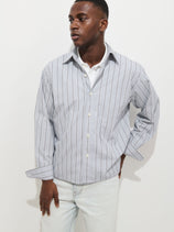 A man stands against a white background wearing the Franklin Striped Shirt in Cotton Poplin, featuring light blue and white stripes over a white tee, paired with light wash jeans. He looks to the side with one hand on his hip.