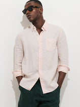 A man stands against a white background, hands in pockets, wearing black sunglasses, dark green pants, and the Mill Shirt In Linen—100% linen with real shell buttons and rolled-up sleeves for a relaxed look.