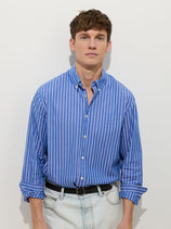 A young man with short brown hair wears the Mill Shirt In Thin Stripe— a 100% cotton blue and white vertical striped button-down — paired with light-wash jeans and a black belt, standing against a plain white background.