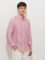 A man with short brown hair stands against a white background, wearing the Mill Striped Shirt In Linen—red and white stripes, real shell buttons, sleeves rolled up—and off-white pants, with his left hand in his pocket.