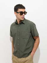 A man wearing black sunglasses, tan pants, and the Franklin Short Sleeve Shirt in Paper Poplin (olive green) stands against a plain white background, looking slightly to his left with his hands in his pockets.