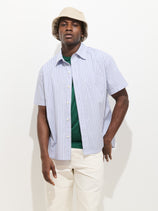 A man wears the Franklin Striped Short Sleeve Shirt in Paper Poplin over a green tee, paired with off-white pants and a beige bucket hat, standing against a plain white background.
