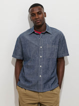 A man stands against a white background wearing the Franklin Short Sleeve Shirt in Chambray, featuring a boxy fit, chest pocket, and short sleeves, paired with tan pants. His hands are in his pockets and he has a neutral expression.