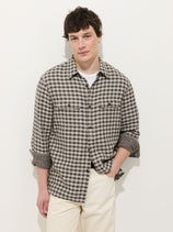 A young man stands against a white background, wearing the Mill Double Gauze Shirt in Cotton Plaid over a white T-shirt and light pants, one hand in his pocket and looking relaxed.