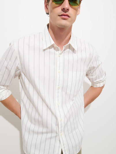 A person wearing the Easy Striped Shirt—a white, long-sleeve, button-up relaxed fit shirt with thin vertical stripes—and green sunglasses stands against a plain white background with hands on hips.