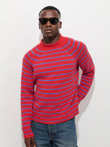 A person wearing the Alex Striped Rollneck In Cotton, featuring red and blue stripes, along with black sunglasses and blue jeans, stands against a plain white background with one hand in their pocket.