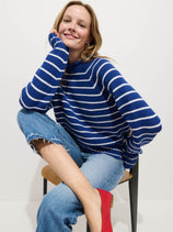 A smiling woman sits on a chair against a white background, wearing the Alex Striped Rollneck In Cotton, ripped blue jeans, and red shoes. One leg is up on the chair as she rests her chin thoughtfully on her hand.