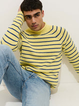 A man with short dark hair and a neutral expression sits against a white wall, wearing the Alex Striped Rollneck In Cotton—a yellow 100% cotton sweater with navy blue horizontal stripes—and light blue jeans.