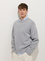 A young man with short brown hair wears the Gabriel Crewneck Sweater in Cotton Linen, layered over a collared shirt and paired with off-white pants, standing against a plain white background and looking at the camera with a neutral expression.