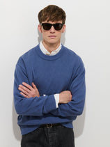 A man stands against a plain white background with his arms crossed, wearing dark sunglasses, black jeans, and the Gabriel Crewneck Sweater in Cotton Linen layered over a white collared shirt.