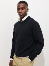 A man with short hair and a neutral expression stands against a white background, wearing the Gabriel Crewneck Sweater in cotton linen (dark navy) over a white collared shirt and beige pants, with one hand tucked into his pocket.