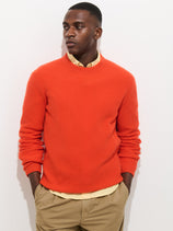 A man wears the Jordan Sweater in lightweight cashmere, bright orange, over a yellow collared shirt and khaki pants, standing against a white background with hands in his pockets, looking to the side.