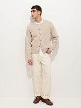 A man with short dark hair stands against a white background wearing the Aaron Cardigan in Cotton Linen over a white shirt, off-white pants, and brown shoes. He looks to the side with one hand in his pocket.