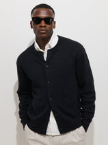 A man in dark sunglasses, a white collared shirt, and the Aaron Marled Cardigan in Cotton Linen stands against a plain white background with his hands in his pockets.