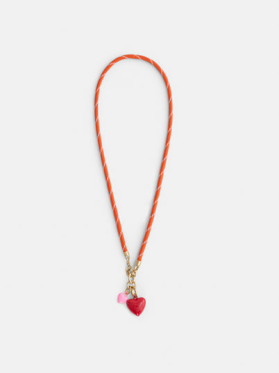 The Roxanne Assoulin Happy Cord Charm Necklace features a puffy heart pendant on a vibrant orange cord with a gold clasp and two removable charms—a red heart and a smaller pink heart—shown on a white background.