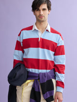 A man with dark hair wears a red and light blue J.Press x Alex Mill Rugby Striped Shirt in cotton and cream pants, standing against a purple background while holding a navy jacket with a purple and black sweater tied around his waist.