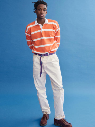 A man stands against a blue background in the J.Press x Alex Mill Rugby Striped Shirt In Cotton, featuring orange and white stripes. He pairs it with cream pants, a purple belt, brown shoes, and looks slightly to the side.