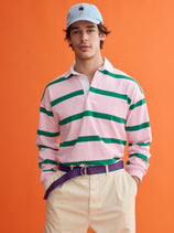 A man in a light blue cap, beige pants, striped belt, and the J.Press x Alex Mill Rugby Striped Shirt in Cotton—pink and green stripes on heavyweight jersey—stands against an orange background.