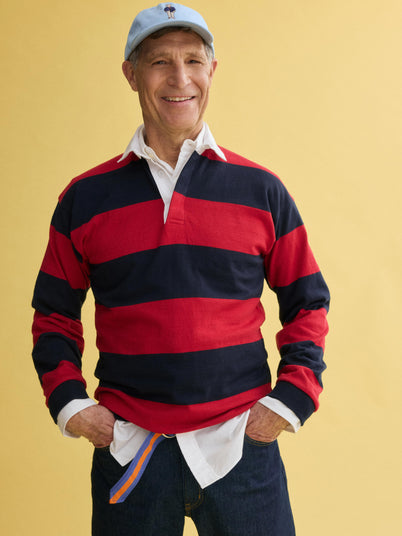 An older man stands smiling with hands in pockets against a yellow background, wearing a J.Press x Alex Mill Rugby Striped Shirt in cotton over a white collared shirt, dark jeans, and a light blue baseball cap.
