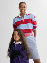 An adult and child smile together in J.Press x Alex Mill Rugby Striped Shirt In Cotton—one wears red and blue stripes with a light blue skirt, the other navy and purple stripes. Both have dark hair and white collars.
