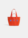 The Painted Perfect Daily Tote is a bright orange bag with two handles, bold blue and white NYC lettering on the front, and metal snap details. Displayed against a white background.