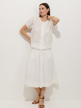 A woman poses against a white background in the Josephine Skirt in ramie—a fully lined, loose, mid-length white skirt with a pull-on waistband—paired with a short-sleeved ruffled blouse, brown sandals, and gold jewelry. Her hand rests on her head.