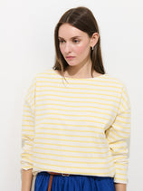 A woman with long brown hair wears the Avignon Striped Tee In Cotton in yellow and white, paired with a blue skirt, standing against a plain white background while looking slightly downward.