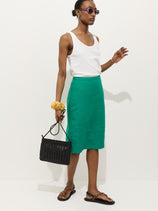 A woman in a white tank top, Maya Pencil Skirt In Linen (green), brown sandals, and purple sunglasses holds a black woven handbag and yellow scrunchie on her wrist, standing against a plain white background.