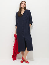 A woman in the Kerry Dress In Linen—a long, navy blue 100% linen button-up—stands against a white background. She holds a red garment and wears red flats, her dark hair loose for a neutral, spring-ready look.