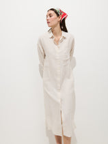 A woman models the Kerry Dress In Linen—a long, light beige 100% linen button-down shirt dress—paired with a colorful headscarf against a plain white background for an effortless spring look.