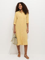 A woman smiles against a white background in the Kerry Dress In Linen, a long-sleeved, button-down yellow dress made from 100% linen. She pairs it with tan sandals, curly hair, and a straw bag featuring a patterned scarf—spring ready!.
