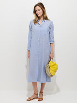 A woman poses against a white backdrop in the Kerry Shirtdress In Seersucker—its loose blue and white stripes paired with brown sandals, a straw handbag, and a yellow cap for an effortlessly chic spring look.