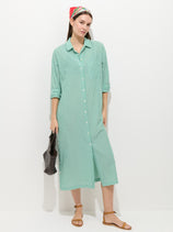 A woman stands against a white background in the Kerry Shirtdress In Seersucker, featuring light green and white stripes. She pairs it with brown sandals, a colorful headscarf, and a woven dark bag—an effortless spring look with rolled sleeves.