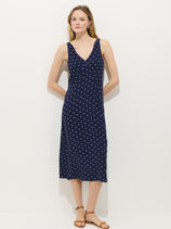 A woman with light brown hair and minimal makeup wears the Flora Dress In Polka Dot—a navy blue, sleeveless, V-neck, bias-cut dress with white polka dots—paired with tan flat sandals against a white background.