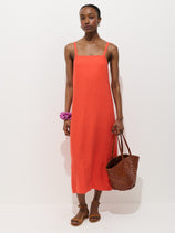 A woman stands against a white background wearing the Marie Dress In Linen, a sleeveless bright orange dress with adjustable straps, styled with brown sandals, a purple scrunchie on her wrist, and carrying a large woven brown bag.