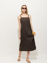 A woman wears the Marie Dress In Linen, a sleeveless dark brown dress with adjustable straps and a square neck, styled with sandals, dark sunglasses, and a chunky bracelet while standing against a plain white background.