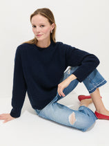 A woman with light skin and straight, light brown hair sits on the floor against a white background. She wears the Charlie Cable Sweater in Cashmere (dark blue), ripped light blue jeans, and red flats, with one arm resting on her knee.