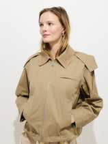 A woman with light brown hair wears the tan Kenzie Jacket, a lightweight water repellent design with a corduroy collar. She stands against a white background, hands in her pockets, gazing slightly upward and to the side.