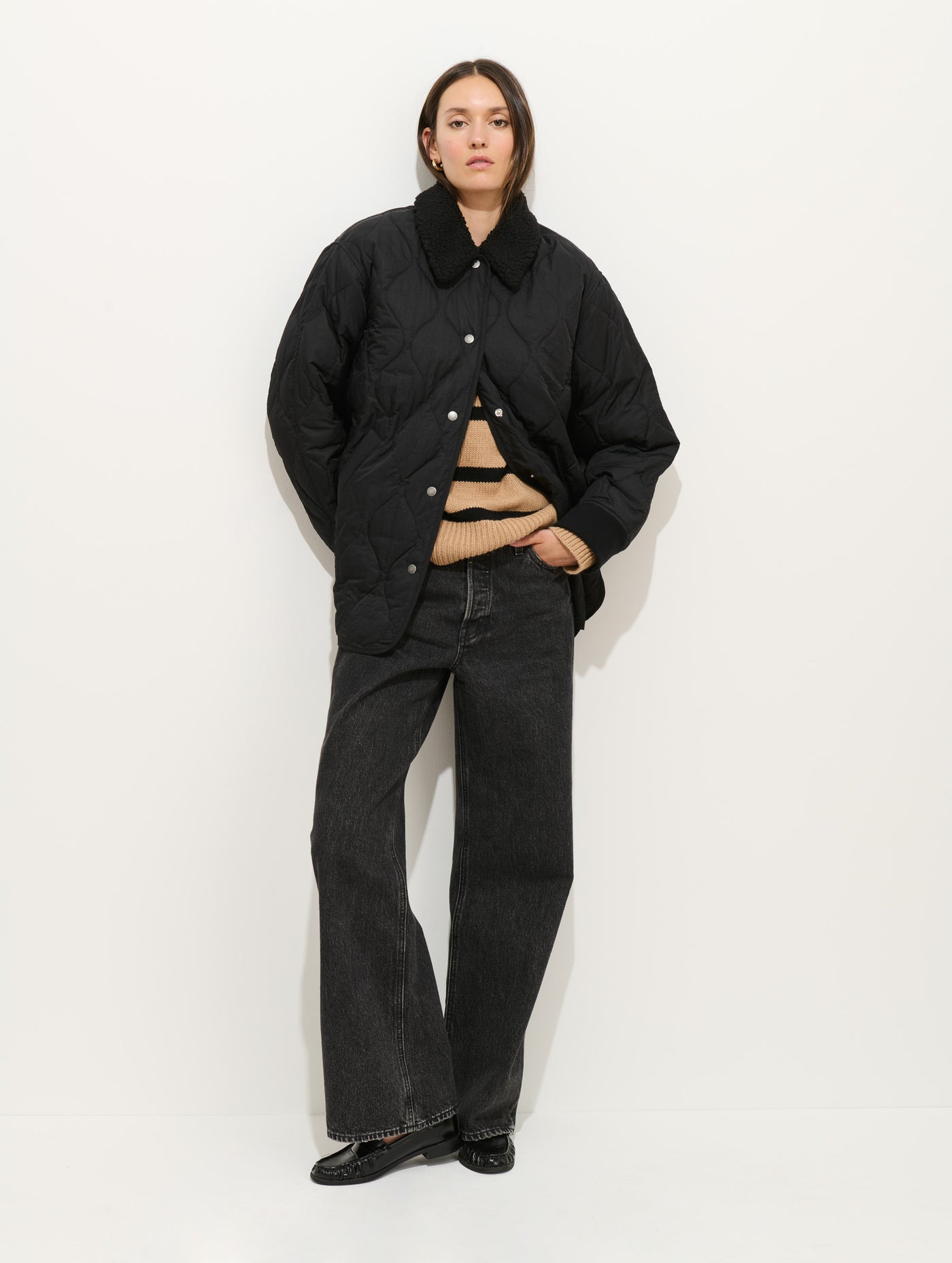 A woman poses against a white background wearing the Wembley Quilted Jacket—black and quilted with a relaxed fit—paired with a tan striped sweater, dark wide-leg jeans, and black shoes. She stands with her hands in her pockets, looking ahead.