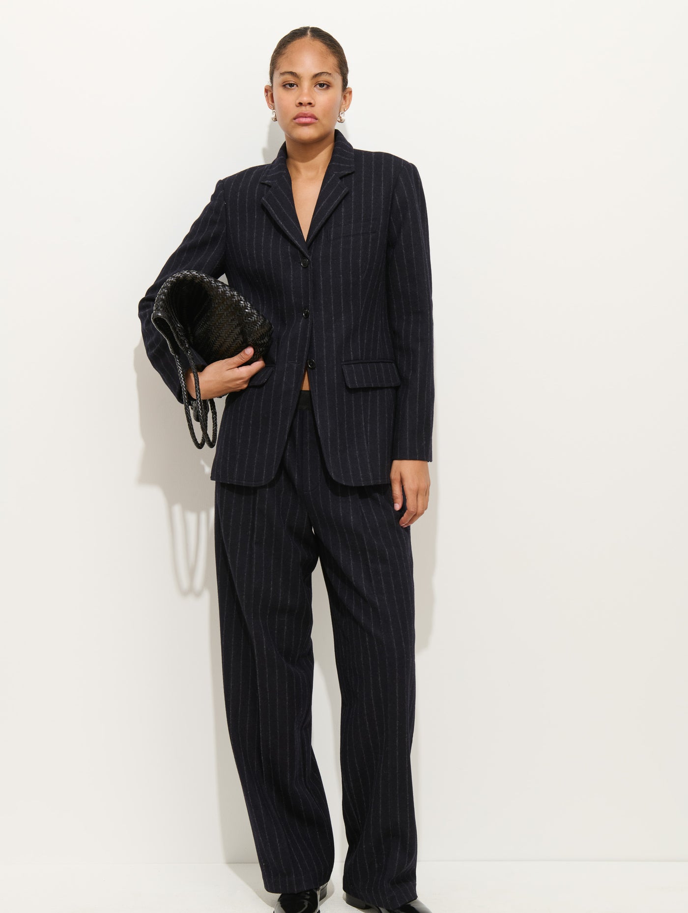 A woman stands against a white background, wearing the Primrose Pleated Trouser in wool with a matching dark pinstriped blazer. She holds a large black woven clutch and looks confidently at the camera.