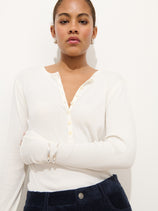 A woman poses against a white background wearing the Laine Henley In Fine Rib and dark blue corduroy pants, styled with hoop earrings and a silver bangle, her expression confident and neutral.