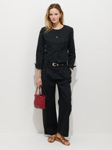 A woman with wavy blonde hair stands against a white wall, wearing a black Soho Jacket and matching Soho Pleated Pant In Cotton Linen, tan sandals, and holding a textured red handbag. She has a neutral expression.