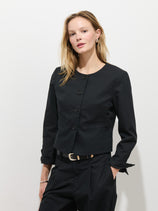 A woman with long blonde hair wears the Soho Jacket In Cotton Linen in black with Corozo nut buttons and matching black pants, standing against a white background and looking at the camera with a neutral expression.