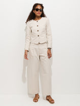 A woman wearing sunglasses, a cream-colored button jacket, Soho Pleated Pant In Cotton Linen, and sandals stands against a plain white background.